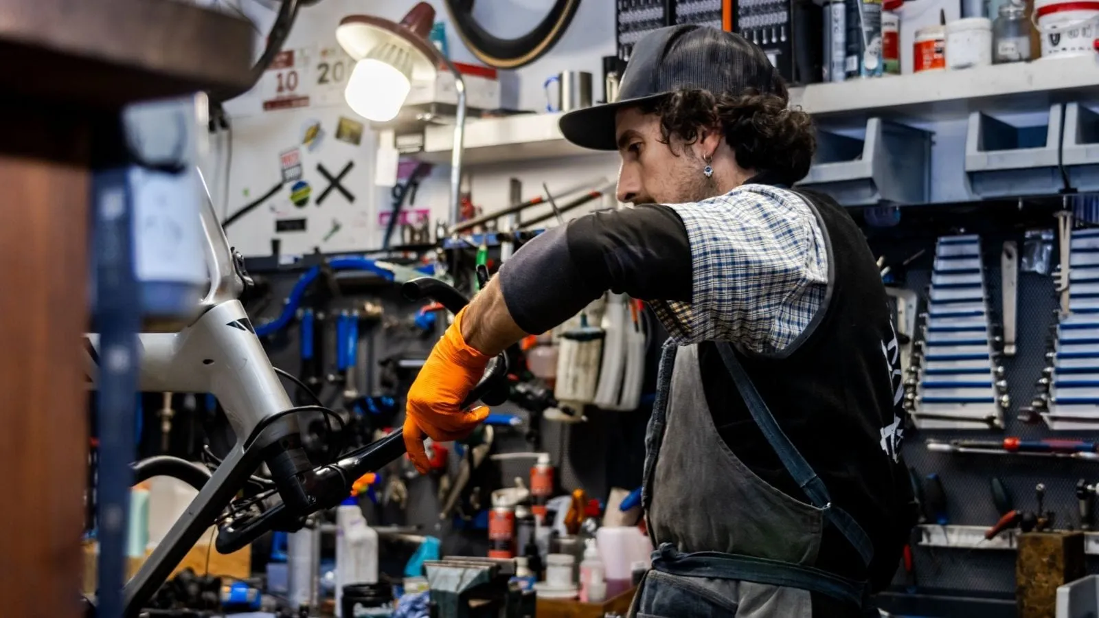 A bike mechanic in a cap and apron working on a bicycle at a tool-covered workbench.